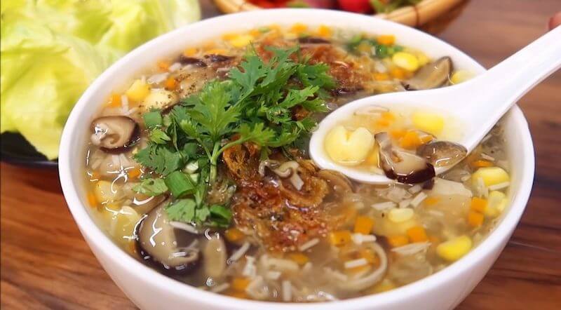 Một tô súp cua, muỗng đang múc súp cua, có nhiều nguyên liệu như nấm, bắp, hành, ngò, nước